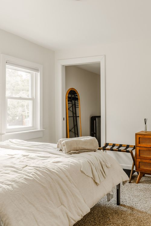 Bedroom corner with a cane nightstand, arched mirror and folded linens on the bed