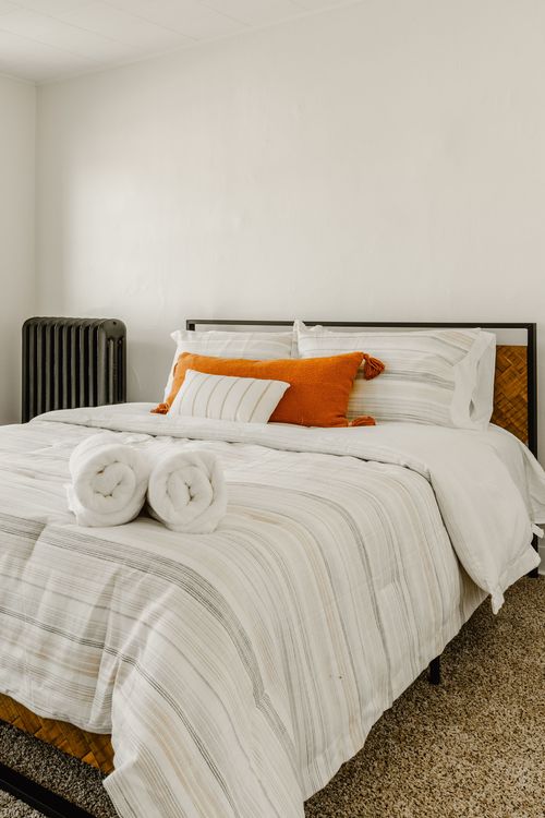 Bedroom with striped white bedding, orange accent pillows and a vintage cast-iron radiator