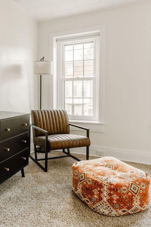 Reading nook with a brown leather armchair, floor lamp and patterned orange pouf by a window