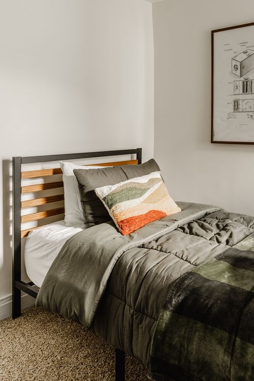 Twin bed with olive green bedding, a mountain-print throw pillow and a framed patent print