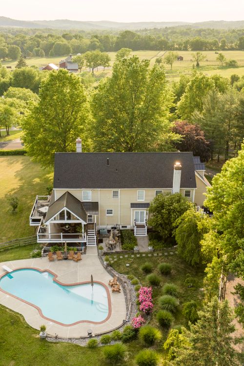 Aerial view of a farmhouse with pool and covered porch overlooking distant pastures and a barn