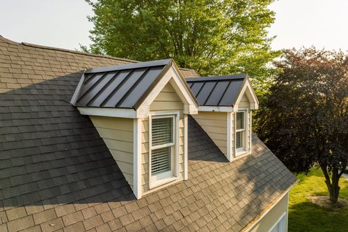 Standing-seam metal dormer roofs set into a shingle roofline with mature trees behind